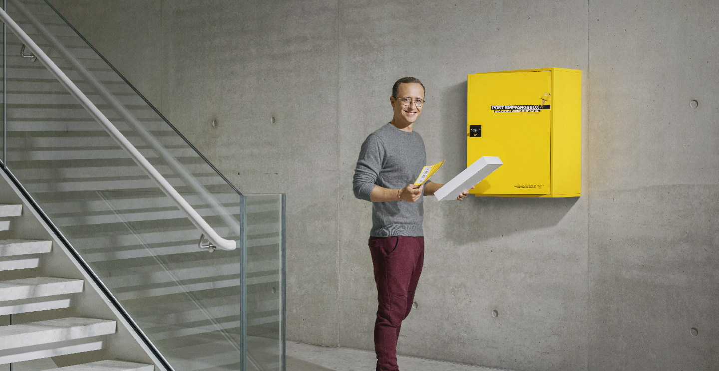 Man standing by a yellow Austrian Post Empfangsbox, holding a package and a letter in a modern building.