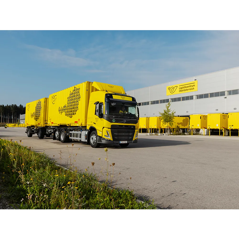 Post-Zustellfahrzeug mit gelben Containern vor dem Logistikzentrum Oberösterreich an einem sonnigen Tag.