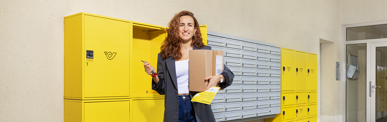 Person holt ein Paket aus gelben Postfächern und hält ein Paket sowie ein Kuvert in der Hand.