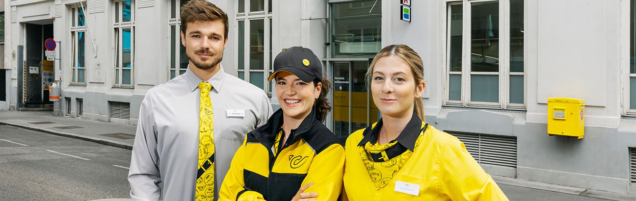 Drei Mitarbeiter der Österreichischen Post in Uniform vor einer Postfiliale mit gelbem Briefkasten an der Wand.