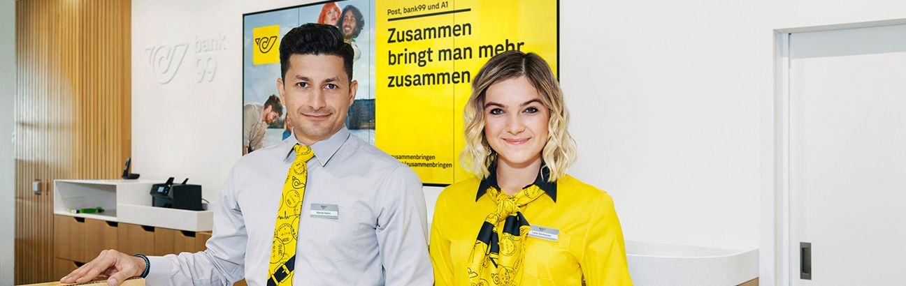 Zwei Mitarbeiter der Österreichischen Post in Uniform am Schalter einer Postfiliale mit gelbem Plakat im Hintergrund.
