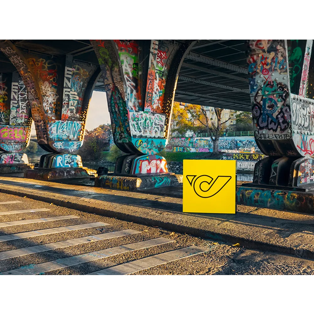 Eine mit Graffiti bedeckte Brücke mit einem gelben Würfel mit Post-Logo im Vordergrund, symbolisiert urbane Präsenz.